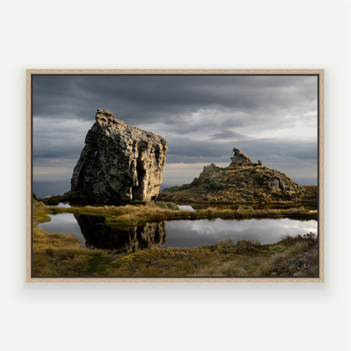 Hump Ridge Track - A framed landscape photograph of a dramatic rocky formation and smaller rock spire reflected in a shallow pool of water under a dark, moody sky, presented in a light wood frame against a white background.