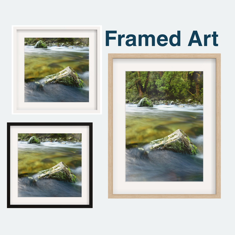 Three framed landscape art prints showing a flowing wild river with mossy rocks, displayed on a light gray background with the text 'Framed Art' above. The prints are shown in white, black, and natural wood frames. By Blankwallnz