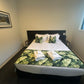 A airbnb with Blankwallnz wall art.
A neatly made bed in a modern bedroom, featuring white linens, green tropical leaf-patterned pillows and runner, two folded white towels, and a dark wood headboard.