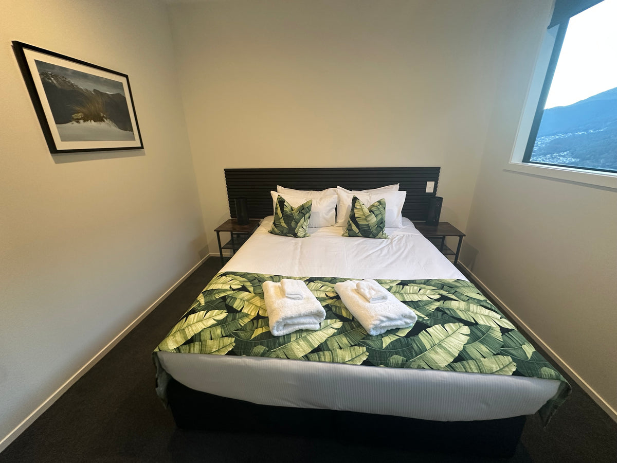 A airbnb with Blankwallnz wall art.
A neatly made bed in a modern bedroom, featuring white linens, green tropical leaf-patterned pillows and runner, two folded white towels, and a dark wood headboard.