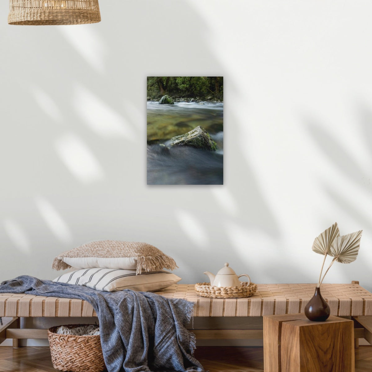 A vertical canvas print of a wild native bush in a national park on New Zealand's Fiordland with mossy rocks, displayed on a white wall above a wooden bench in a styled room setting.
