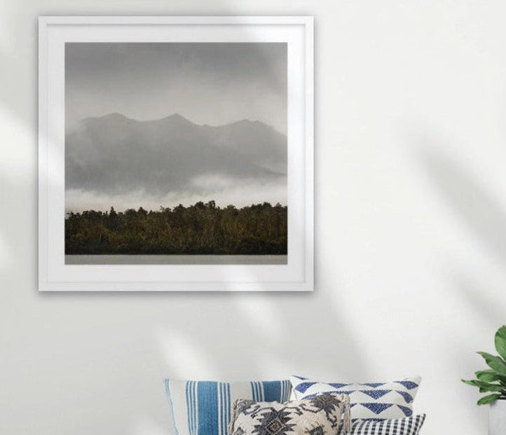 A framed New Zealand moody landscape art print depicting misty mountains and a dark forest, displayed on a white wall above a couch with decorative pillows and a small plant in a home setting.