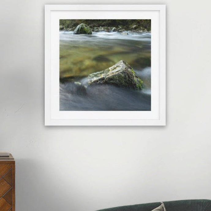 A framed wilderness art print depicting a mossy rock in a flowing river, displayed on a light gray wall above a wooden sideboard and next to a dark green sofa.