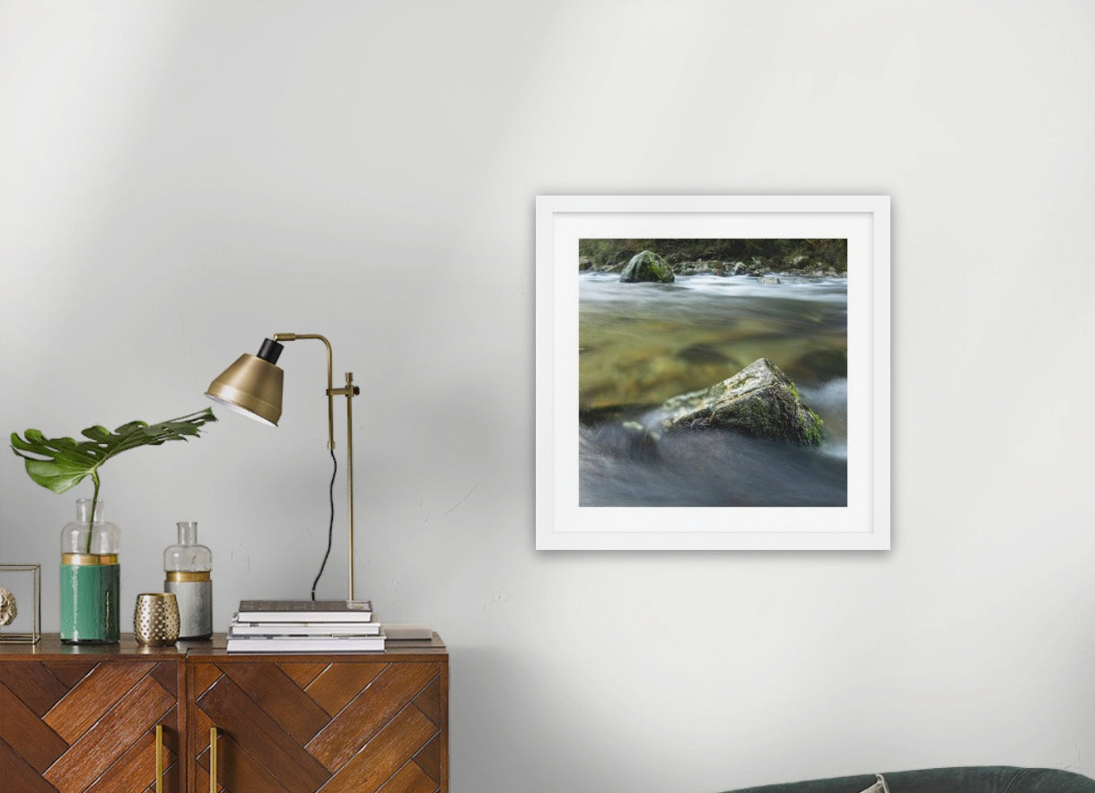 A framed art Wilderness art print depicting a mossy log in a flowing river, styled on a light gray wall above a wooden sideboard .