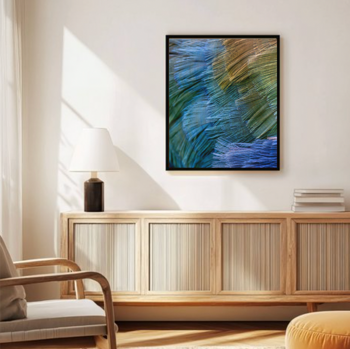 A framed abstract real photo wall art print with blue, green, and gold Takehe feathers hangs above a light wood sideboard in a styled living room with a chair, lamp, books, and a round pouf.