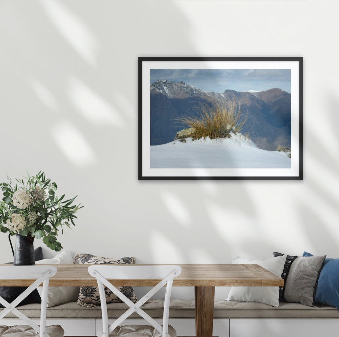 A framed landscape art print of snow-covered mountains with a native snow tussock under a moody sky, displayed on a white wall above a dining table in a modern room setting. Wilderness wall art by Blankwallnz