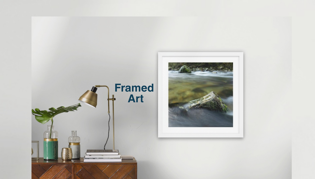 A framed New Zealand wild rover landscape art print depicting a river with a mossy rock, displayed on a light gray wall above a wooden sideboard with a lamp and decorative items. The text 'Framed Art' is visible next to the frame.