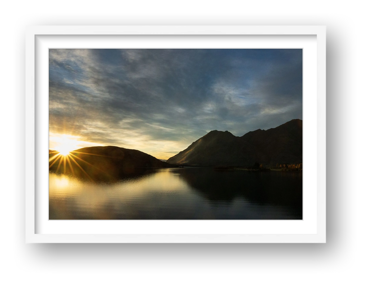 Wanaka Break of Day peaks over the isthmus on to Glendhu Bay on Lake Wānaka. Streaky skies and dark hills give hints of an oil painting. Blue yellow dawn.